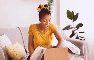 Happy Customer Concept. Portrait of excited black woman opening and unpacking cardboard box parcel, sitting on the couch in living room at home. Young happy female satisfied with delivery service photo