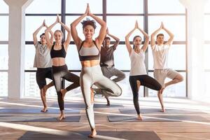 Wellness Concept. Group Of Sporty People Practicing Yoga With Instructor In Class, Standing In Tree Pose Against Window In Loft Studio photo