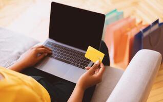 Online Shopping And Payment. Over the shoulder view of african american woman holding credit card in hand and using laptop with black blank screen, typing on keyboard, sitting on couch, copy space photo