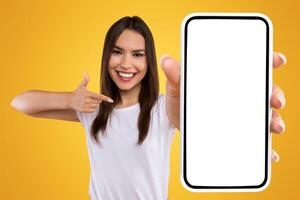Check This Website. Portrait of smiling female holding smartphone with white blank screen in hand, showing device close up to camera and pointing finger at gadget with empty free space for mock up photo