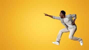 Crazy Sales. Emotional black man pointing aside while jumping in air over beige studio background, overjoyed excited african american guy showing copy space for design or advertisement, panorama photo