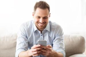 Messaging At Social Network. Happy millennial guy using modern smartphone device, sitting on sofa at home photo