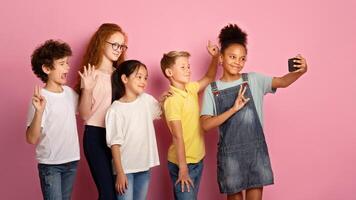 Multinational schoolkids taking selfie together on pink background photo