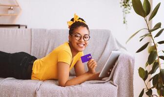 Easy Online Payment Concept. Smiling black lady in glasses using digital tablet and showing credit card, ordering purchases via internet. Woman lying on the couch and looking at camera photo