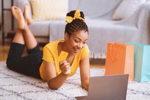 Online Shopping Concept. Excited african american lady showing credit card and using laptop, ordering clothes and purchases via internet, lying on the floor carpet in living room at home photo