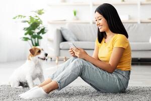 Smiling young asian lady sitting on floor at home, checking photos of her cute puppy, using cellphone, side view, copy space, jack russel terrier owner watching educational on Internet