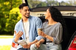 Happy multiracial millennial couple having break at countryside while car trip, sitting on open auto trunk, drinking coffee and having conversation, discussing details of their journey, free space photo