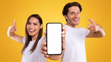 Use this New Application. Portrait of cheerful millennial couple pointing at smartphone with white screen, recommending mobile app, advertising your website, mock up for design. Selective focus photo