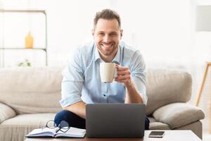 Good Morning. Smiling man drinking coffee at home and using laptop, looking at camera, copyspace photo