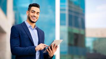 Good-looking arab guy in formal wear standing by office building, holding digital tablet and smiling, looking for new business opportunities online, using business application, copy space, panorama photo