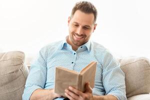 Time To Read. Smiling man holding paper book, resting on couch at home, copy space photo