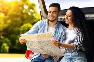 Loving middle-eastern couple watching road map while having break, travelling by car together, copy space. Smiling arab man and pretty brunette lady sitting on open auto trunk, checking location photo