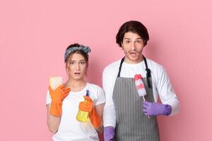 Young Couple With Cleaning Tools In Hands Looking With Shock At Camera, Doesn't Know Where To Start Tidying, Standing On Pink Background, Empty Space photo