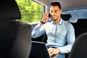 Happy middle-eastern businessman in suit sitting in car, using laptop and having business conversation on smartphone, working online while going to airport, copy space. Modern technologies in business photo