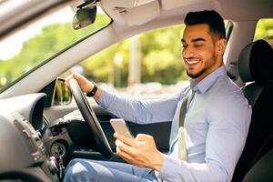 Bad driving habits, unsafe driving, driving and gadgets concept. Cheerful arab man entrepreneur going to office by auto, using cellphone while driving car, chatting with someone, side view, copy space photo