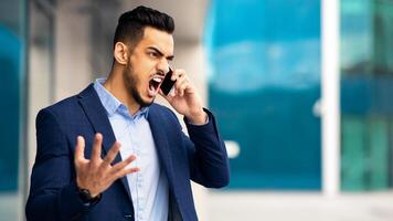 Furious young arab man in suit having phone conversation with assistant or business partner, screaming and gesturing while standing on the street next to office building, panorama with copy space photo