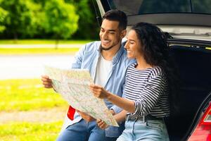 Loving middle-eastern couple watching road map while having break, travelling by car together, panorama with copy space. Smiling arab man and pretty ady sitting on open auto trunk, checking location photo