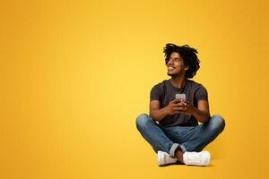 Mobile Offer. Smiling young black man with smartphone in hands sitting on floor and looking aside at copy space on beige studio background, enjoying using modern gadget for communication, panorama photo