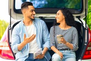 Carefree multiracial millennial couple having break at countryside while car trip, sitting on open trunk, drinking coffee and having conversation, discussing details of their journey, free space photo
