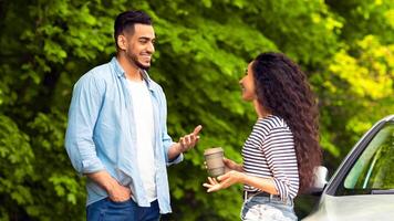 Joyful multiracial millennial couple having coffee break at countryside while car trip, standing by auto, drinking tea and having conversation, discussing details of their journey, free space photo