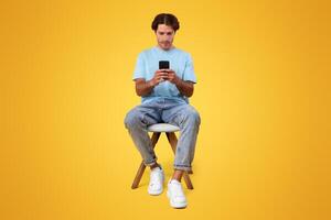 Full Body Length Vertical Portrait Of Focused Millennial Man Using Phone Sitting In Chair Isolated Over Orange Studio Background. Serious Guy Using New Mobile Application On Smartphone, Checking Email photo