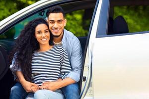 Beautiful middle-eastern lovers enjoying car trip to countryside, closeup portrait. Handsome arab guy hugging his pretty woman, sitting inside open auto, hugging and smiling at camera photo