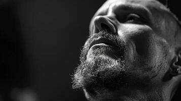 A man with a well groomed beard and intense expression looks upward, illuminated by soft light, highlighting his facial features in a moody atmosphere. photo