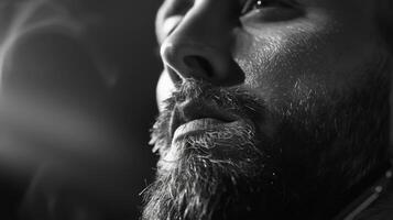 A man with a well groomed beard gazes thoughtfully with a serious expression, illuminated by soft lighting, in a relaxed and artistic environment during the evening. photo