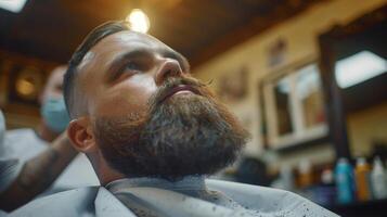 A man with a full beard is getting a trim at a barbershop. He is relaxed in a cape while a barber carefully styles his facial hair. The atmosphere is busy and inviting. photo