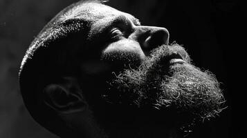 A man with a full beard tilts his head back, evoking a contemplative mood. The interplay of light and shadows creates a striking visual, highlighting facial features. photo