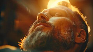 A bearded man relaxes, eyes closed, basking in the warm glow of sunlight. The atmosphere is soothing as he savors a moment of peace and serenity in an inviting indoor space. photo