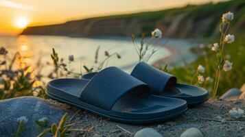 Black sandals rest on pebbles near a sandy beach as the sun sets over calm ocean waves, while gentle greenery adorns the shoreline in the background. photo