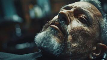 A relaxed mature man lies back in a barbershop chair while receiving a grooming treatment. Subtle reflections of warm lights create an intimate atmosphere. photo