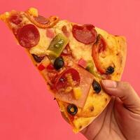hand holding a slice of pizza with toppings like pepperoni, olives, and vegetables against pink background photo