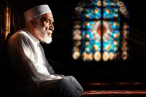 elderly man wearing white robe and kufi cap sits pensively on wooden bench in mosque courtyard. Intricate mosaic tiles create vibrant backdrop as he reflects during festive Eid gatherings photo