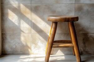 A rustic wooden stool sits in sunlight, casting soft shadows on a textured wall. photo