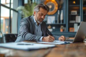 A focused middle-aged Caucasian man in a gray suit writes notes at his desk, with a laptop and modern decor in the background. photo