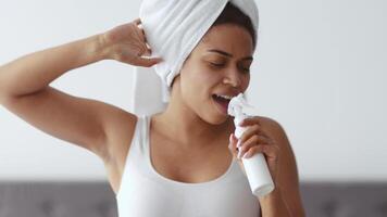 A young African American woman is engaged in her body care routine at home. She appears relaxed, applying a beauty product while wearing a towel on her head, showcasing a moment of self-care. video
