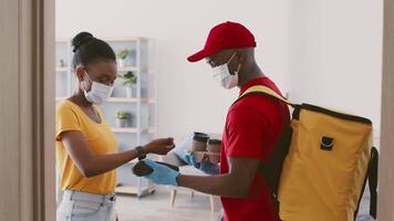 A man in a red cap and yellow backpack delivers a package while wearing gloves and a mask. A woman in a yellow shirt receives the delivery in her apartment with a friendly expression. video