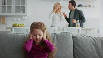 A young girl sits quietly on the couch, looking downcast while her parents argue in the background. The atmosphere is tense, highlighting the impact of domestic conflict on family dynamics. video