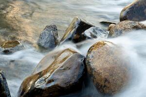 Shows a close-up of a riverbed with smooth rounded stones. Water is flowing gently over the rocks creating a blurred and ethereal effect. photo