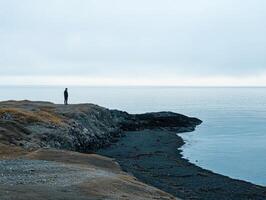 Solitary figure standing on rocky coast with calm waters during overcast afternoon photo