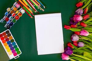 A blank notebook, paint brushes, and a bouquet of tulips on a green table photo