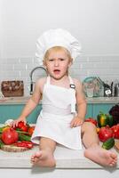 A little girl in a chef's hat sitting on a counter with a bunch of vegetables photo