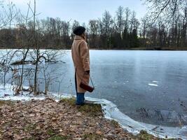 A woman in a warm beige coat and knit hat stands by the edge of a partially frozen lake. The surrounding autumn landscape is dotted with fallen leaves, and bare trees line the horizon under a cool photo