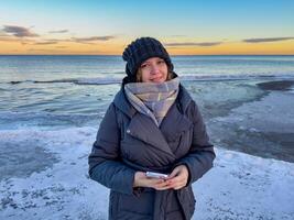 A woman in a warm winter coat, knitted hat, and scarf stands by the frozen shore of Lake Ladoga, smiling gently at the camera. The golden hues of the sunset reflect on the icy water, creating a serene photo