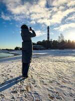 A woman in a long winter coat captures a photo of Osinovets Lighthouse with her smartphone while standing on a snow-covered beach of Lake Ladoga. The setting sun casts long shadows, highlighting the