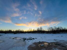 A serene winter sunset paints the sky in shades of blue, orange, and gold over a frozen shoreline. Patches of snow and ice cover the ground, reflecting the soft glow of the setting sun. Bare trees photo