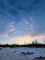 A serene winter sunset paints the sky in shades of blue, orange, and gold over a frozen shoreline. Patches of snow and ice cover the ground, reflecting the soft glow of the setting sun. Bare trees photo