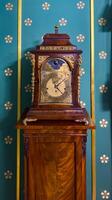 A close-up view of a vintage wooden clock with intricate brass details, set against a rich teal wall adorned with floral patterns. The clocks elegant design reflects the craftsmanship of a bygone era photo